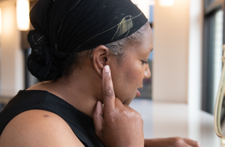 A Black woman tests her hearing aids after putting them on. Her right hand is on her right ear while her left hand holds the hard case for the hearing aids. The woman is wearing black and her hair is pulled back by a scarf.
